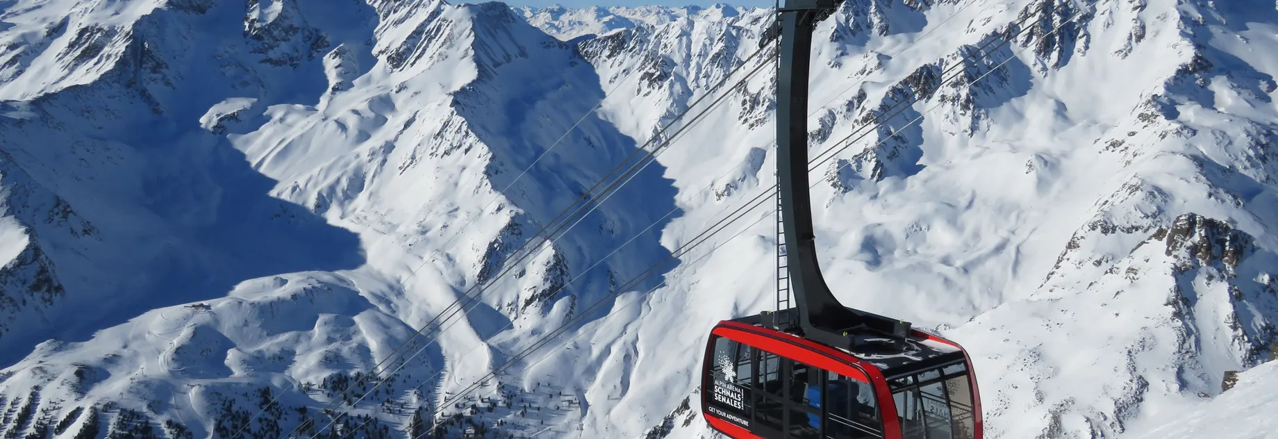 A red cable car travels across snow-covered mountains under a clear blue sky, with rugged peaks and deep valleys in the background.