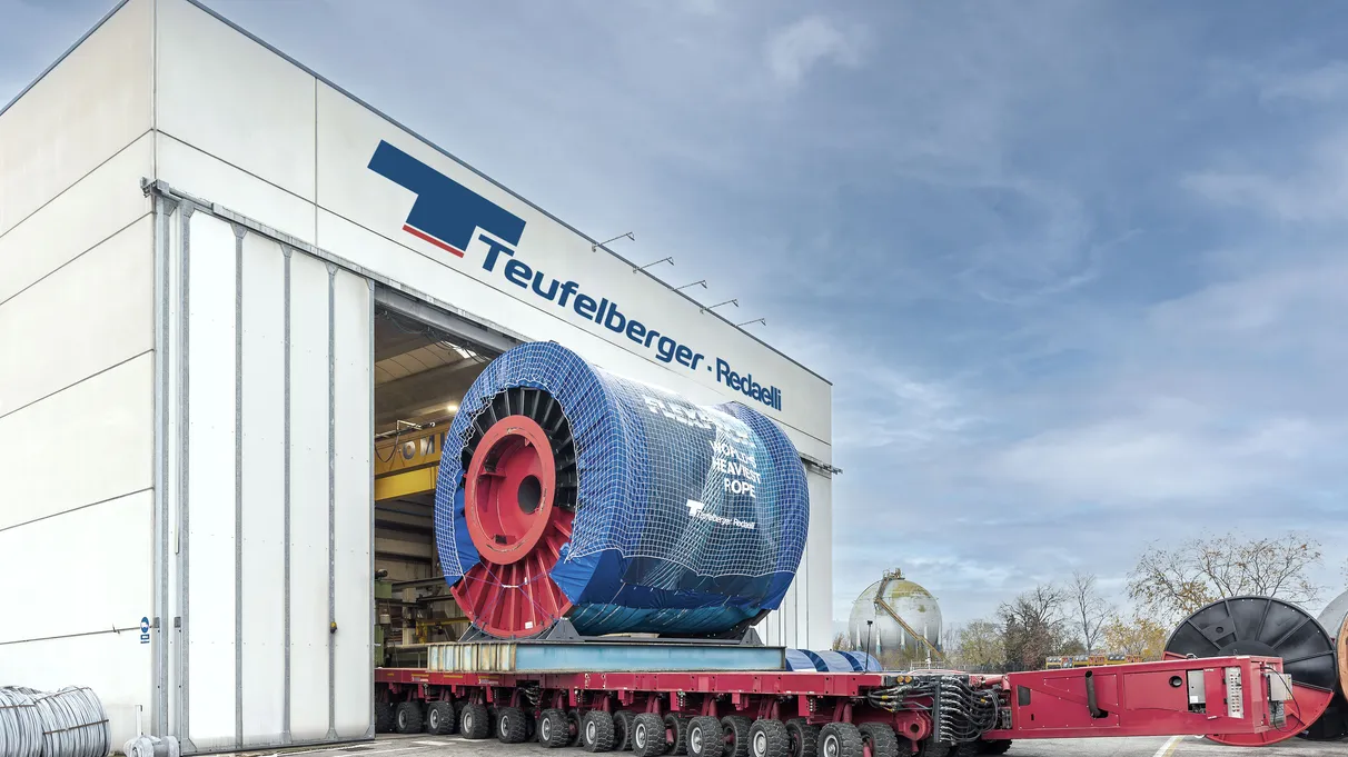 A large industrial cable spool is being transported on a red multi-wheeled trailer from a building labeled "Teufelberger-Redaelli."