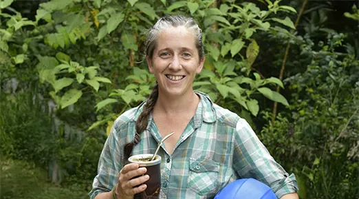 Sofia standing in front of some green bushes holding a cup with a straw. With the left hand she is holding a blue helmet.