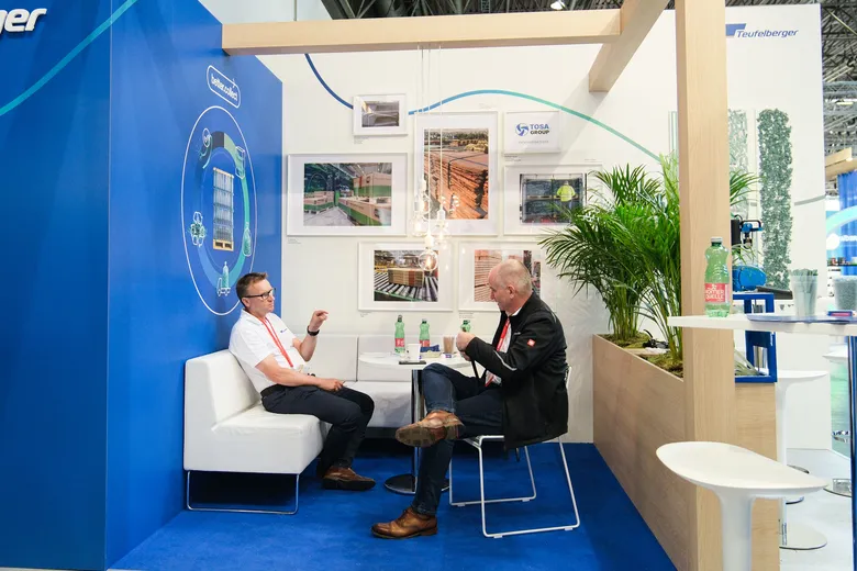 Two men having a discussion at a trade fair booth with seating, blue backdrop, and framed images, emphasizing professional interaction.