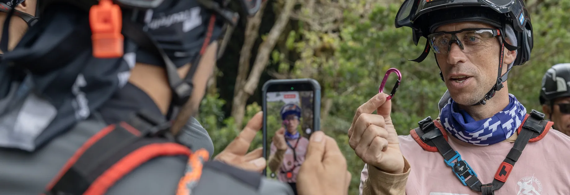 Two helmeted individuals in outdoor gear, one holding a carabiner and the other filming with a phone. Forest background.