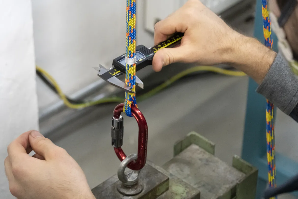 Close-up of hands using a digital caliper to measure the diameter of a colorful climbing rope threaded through a red carabiner, in an industrial setting with equipment in the background.