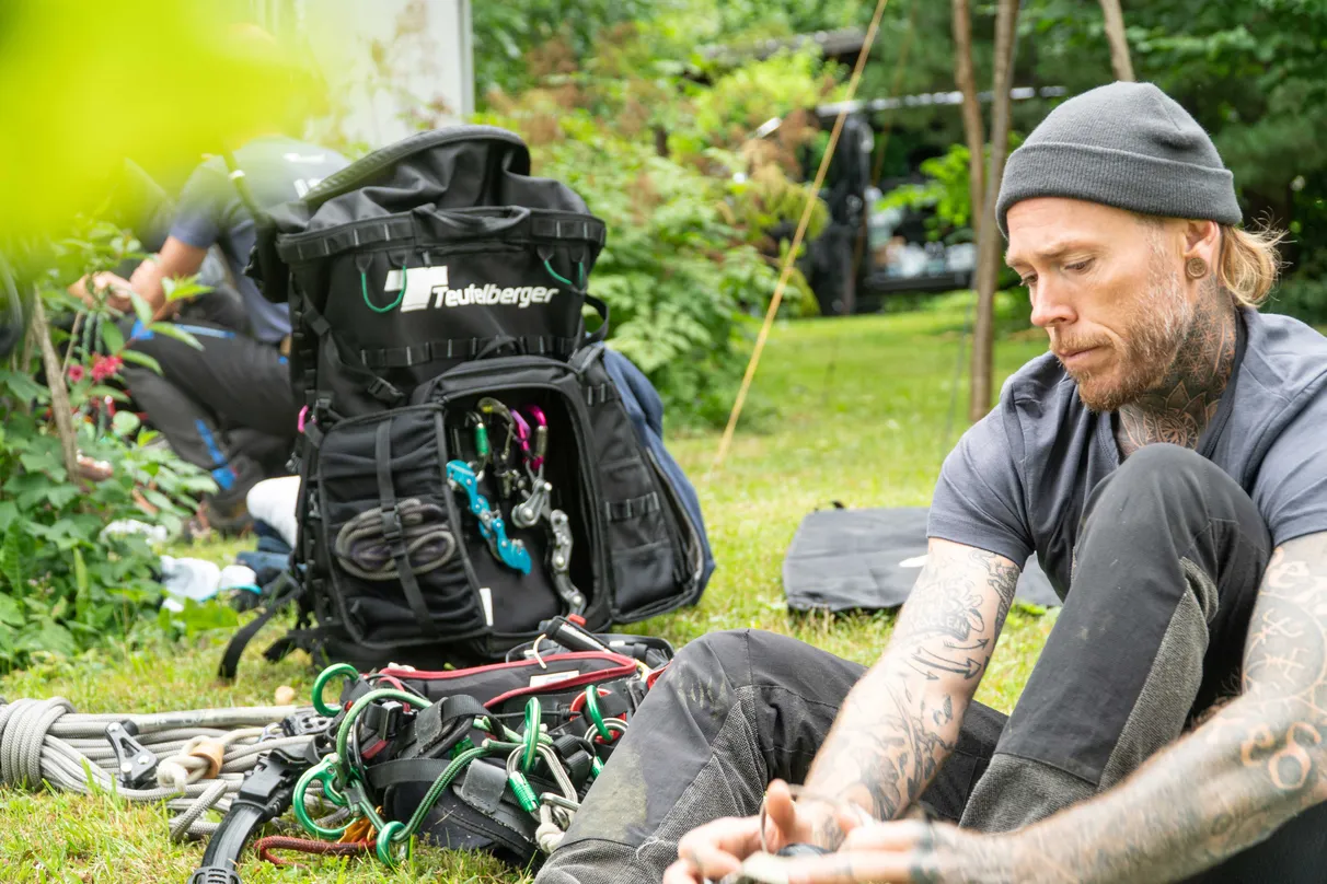 Man sitting on grass with climbing gear and a backpack nearby, focused on preparing equipment.
