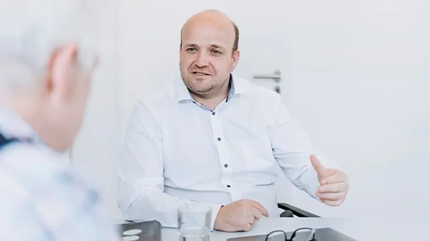 A man in a white shirt sitting at a table, speaking and gesturing with his hand, engaged in conversation with another person in the foreground.