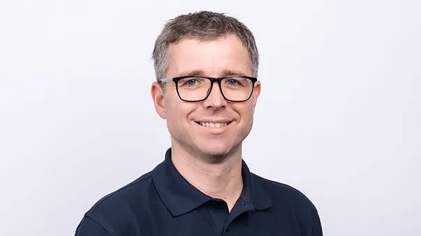 A man wearing glasses and a dark suit jacket over a white shirt, looking at the camera against a plain background.