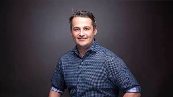 A man wearing a checkered shirt smiles against a dark background. Hair is neatly styled, and he is posing confidently.