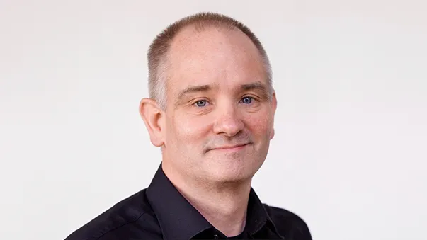 A man with short hair and wearing a black shirt smiles gently against a plain background.