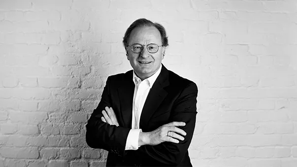 A man in a suit stands confidently with arms crossed against a white brick wall. Black and white image.