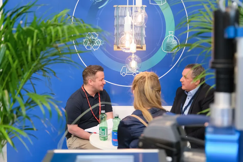 Three people engaged in a professional discussion at a booth, with a blue backdrop and green plants surrounding them.