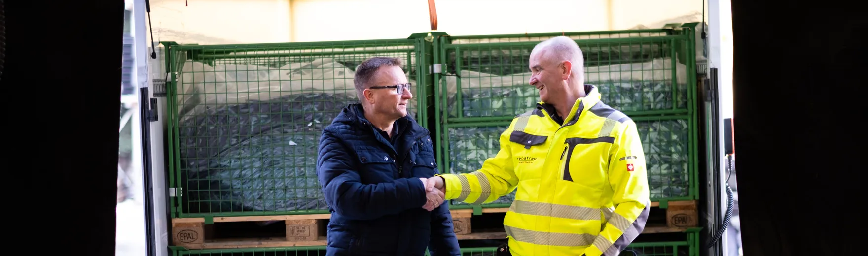 Two men shaking hands in a warehouse setting, with stacked crates in the background, one wearing a high-visibility jacket.