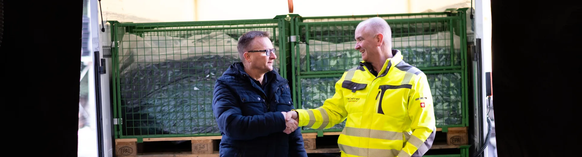 Two men shaking hands in a warehouse setting, with stacked crates in the background, one wearing a high-visibility jacket.