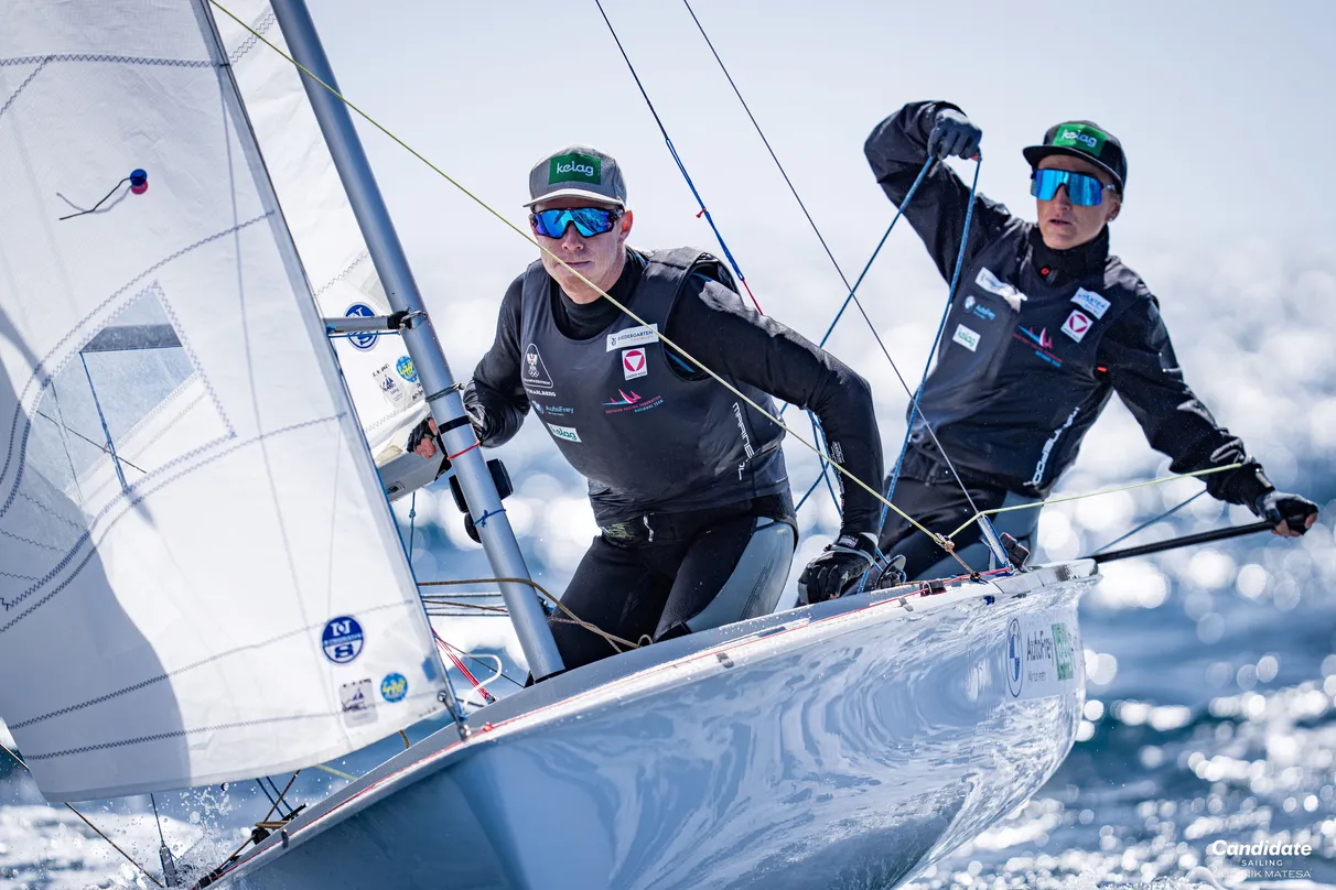 Two sailors in athletic gear maneuver a sailboat on a sunny day, focused and determined, with a clear blue sky and water in the background.