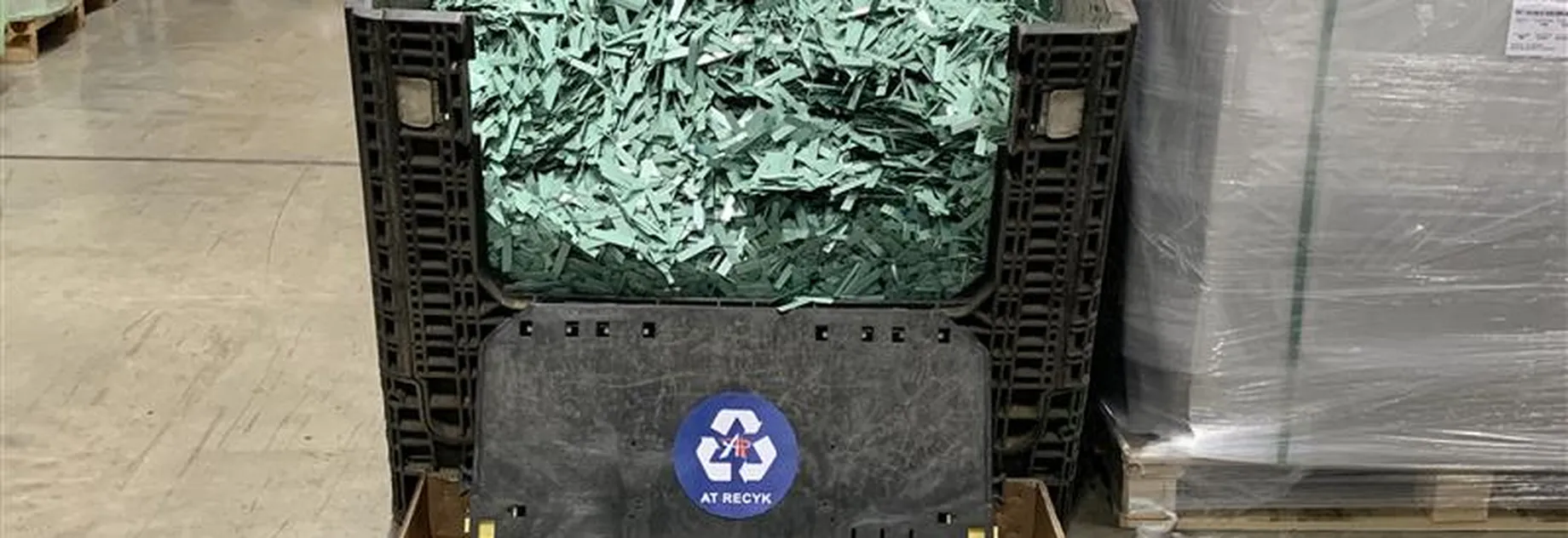 Large bin filled with shredded green material labeled for recycling, situated in an industrial warehouse setting.