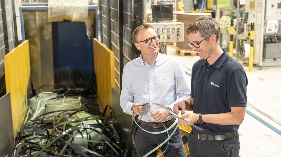 Two men in a factory examine strips of material near a machine. One wears a light shirt and glasses, the other a dark polo shirt.