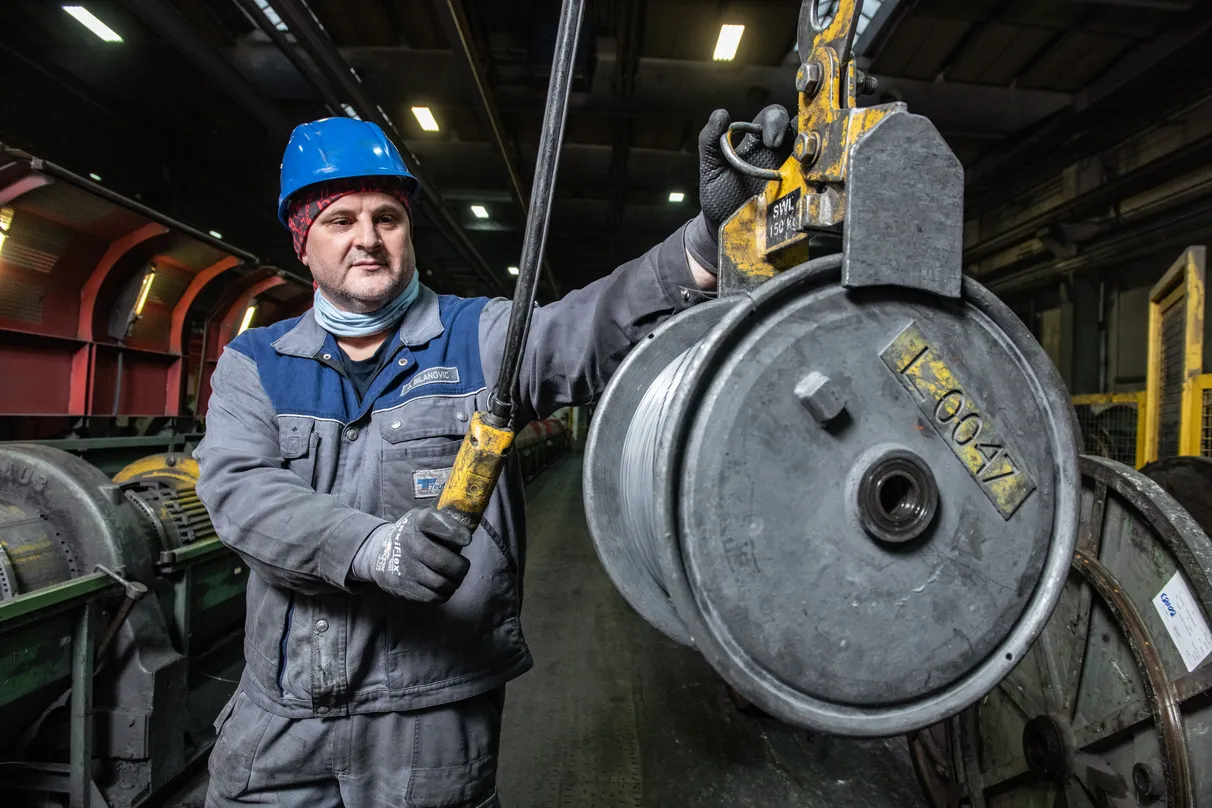 Worker in industrial setting wearing a blue safety helmet and gloves, holding a large machinery part labeled “L7004”. Additional equipment is visible in the background.