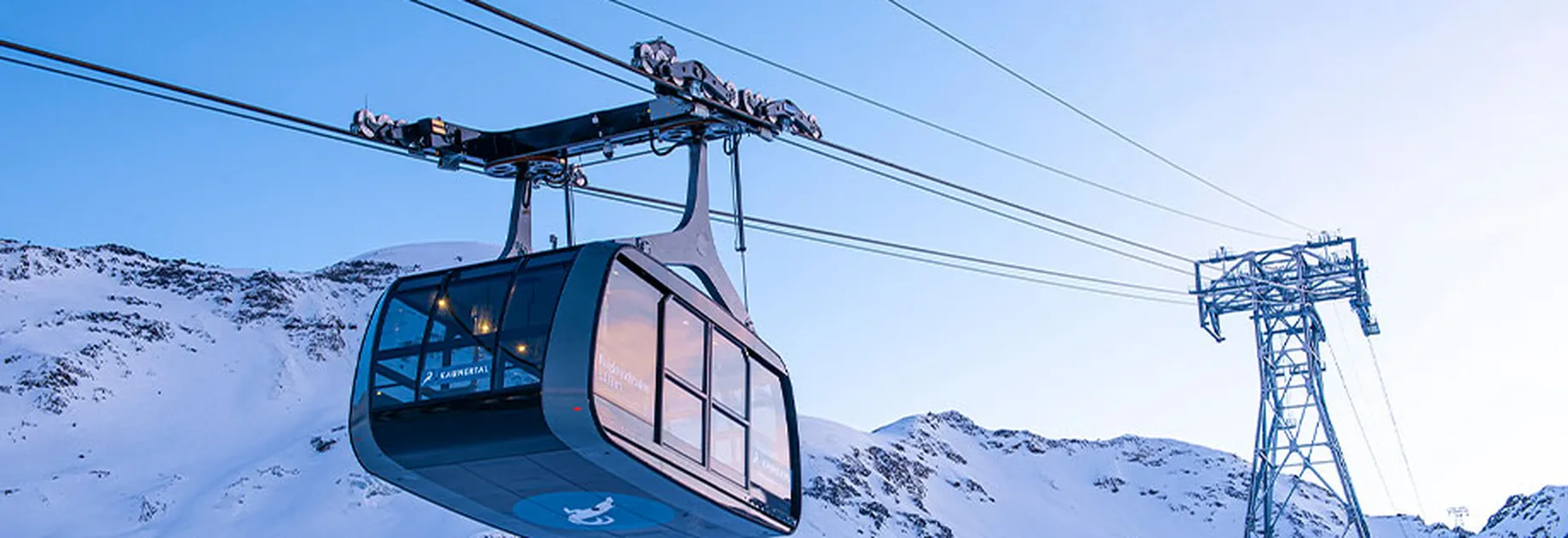 Cable car suspended on wires, traveling over a snowy mountain landscape with a clear blue sky in the background.