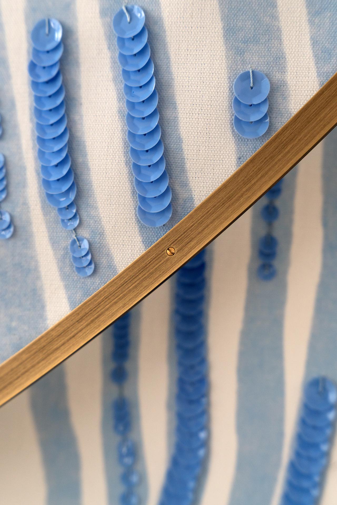 Close-up of blue sequins on striped fabric, with wooden embroidery hoops. Sequins create vertical patterns against light and dark blue stripes.