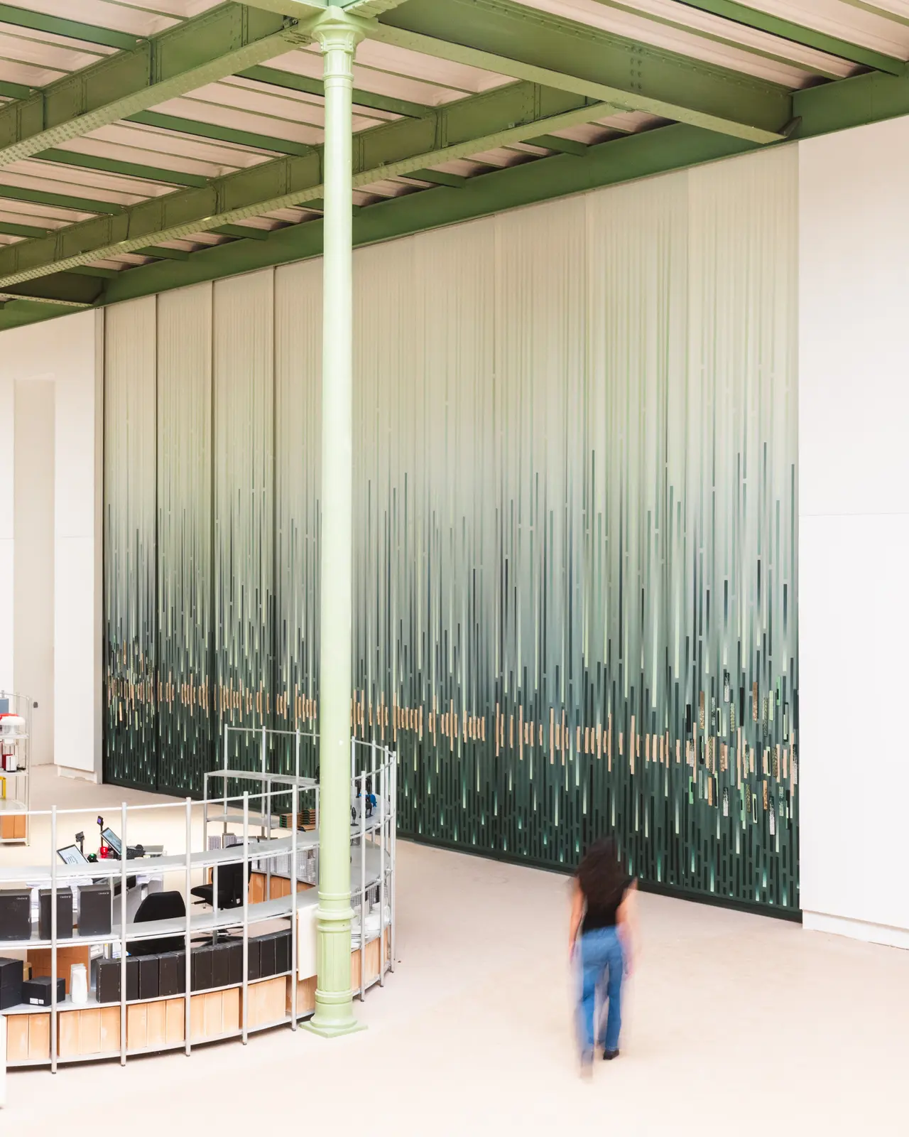 A person walks in a spacious room with a green and white gradient wall design, featuring vertical lines. A circular desk is visible on the left.