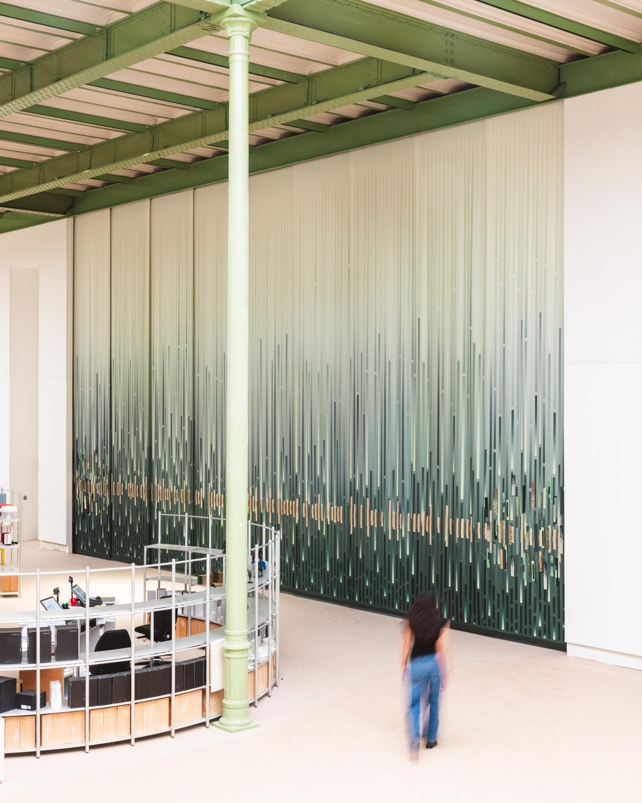 A person walks in a spacious room with a green and white gradient wall design, featuring vertical lines. A circular desk is visible on the left.