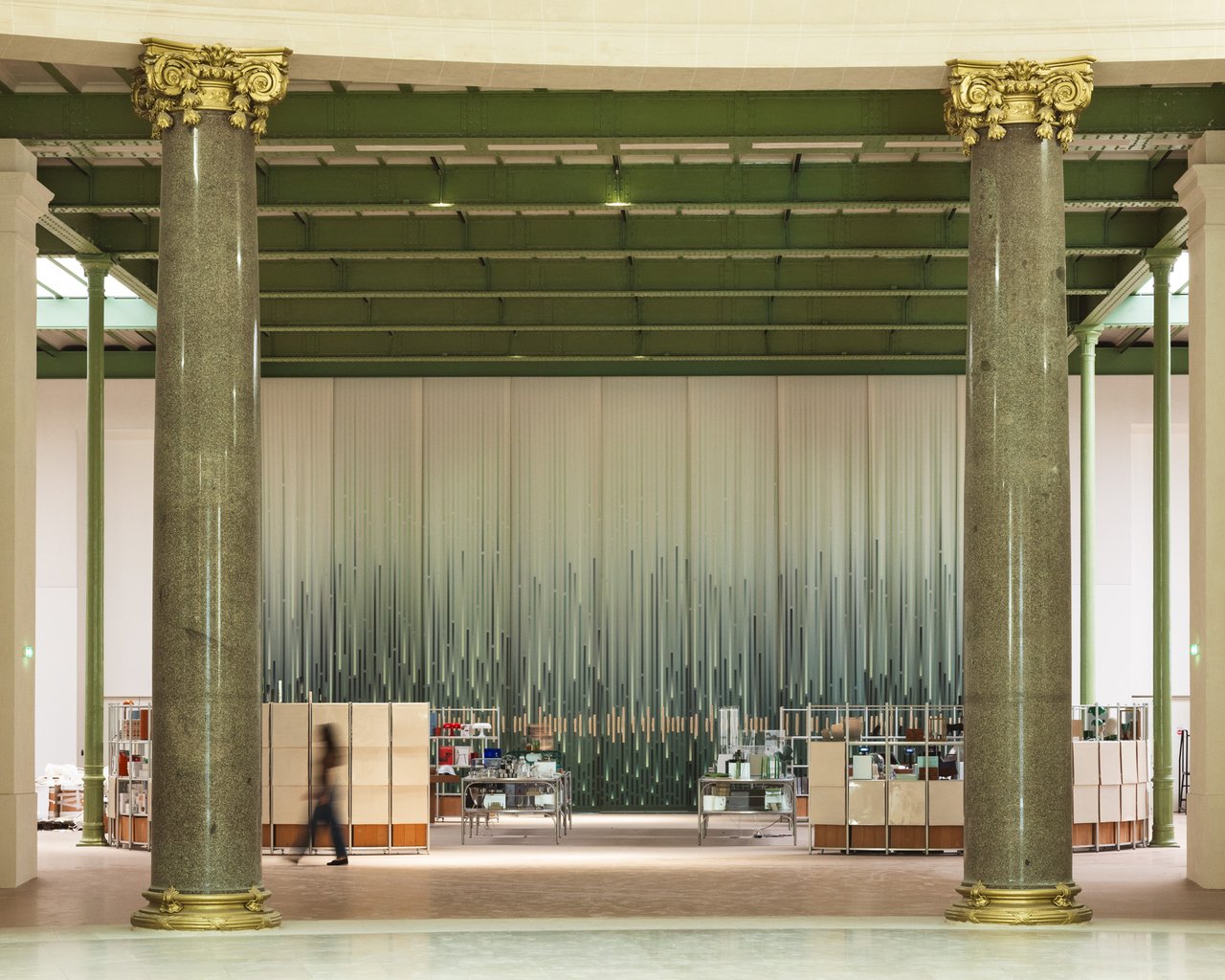 Spacious hall with tall green columns, a modern art installation of vertical lines, and a person walking past shelves of items.