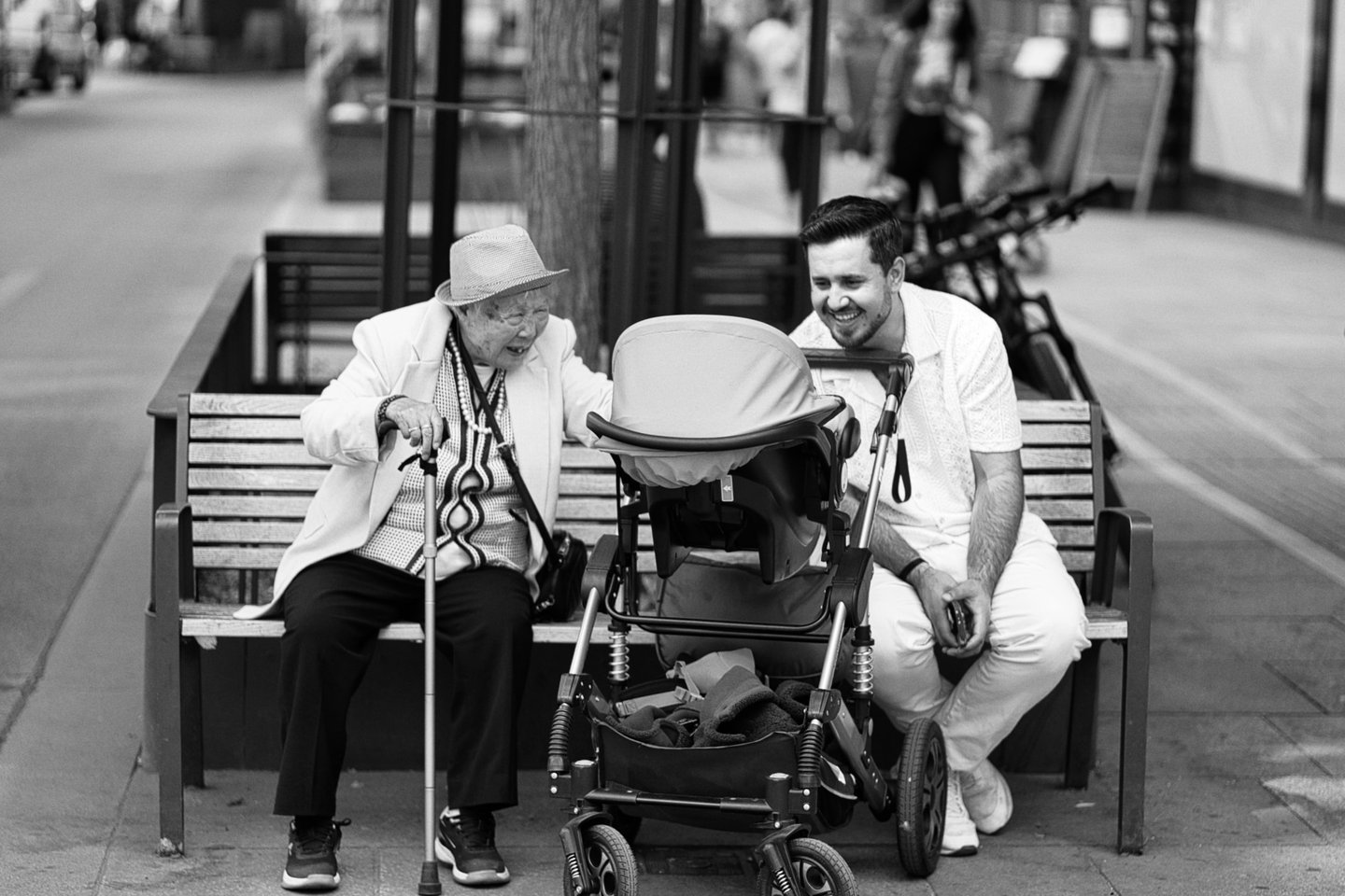 An elderly person and a man sit on a bench, smiling at a baby in a stroller. Sony A7C; Sony FE 85mm f/1.8