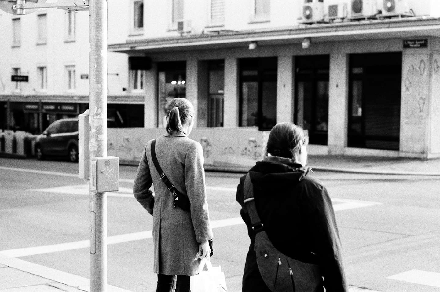 Two women with bags stand at a crosswalk on a city street, shot on film.