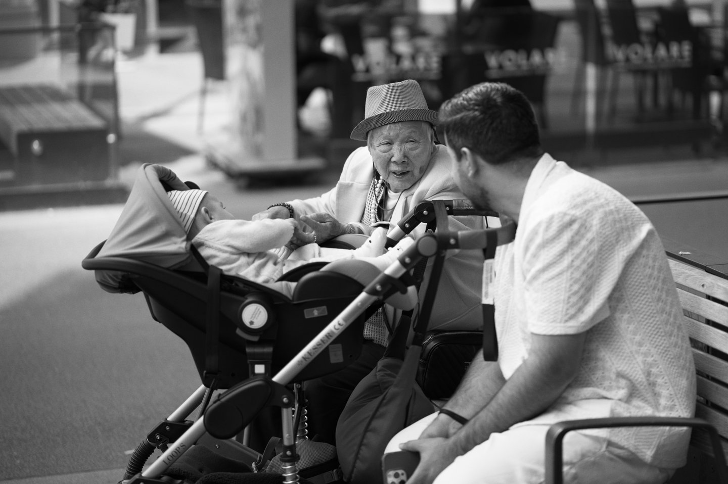 An elderly person and a man sit on a bench, chatting over a baby in a stroller. Sony A7C; Sony FE 85mm f/1.8