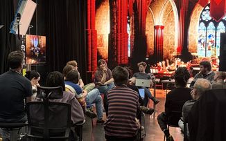 A group of people sit in a circle having a discussion in a studio with gothic-style image projected onto a VP wall and lighting equipment visible.