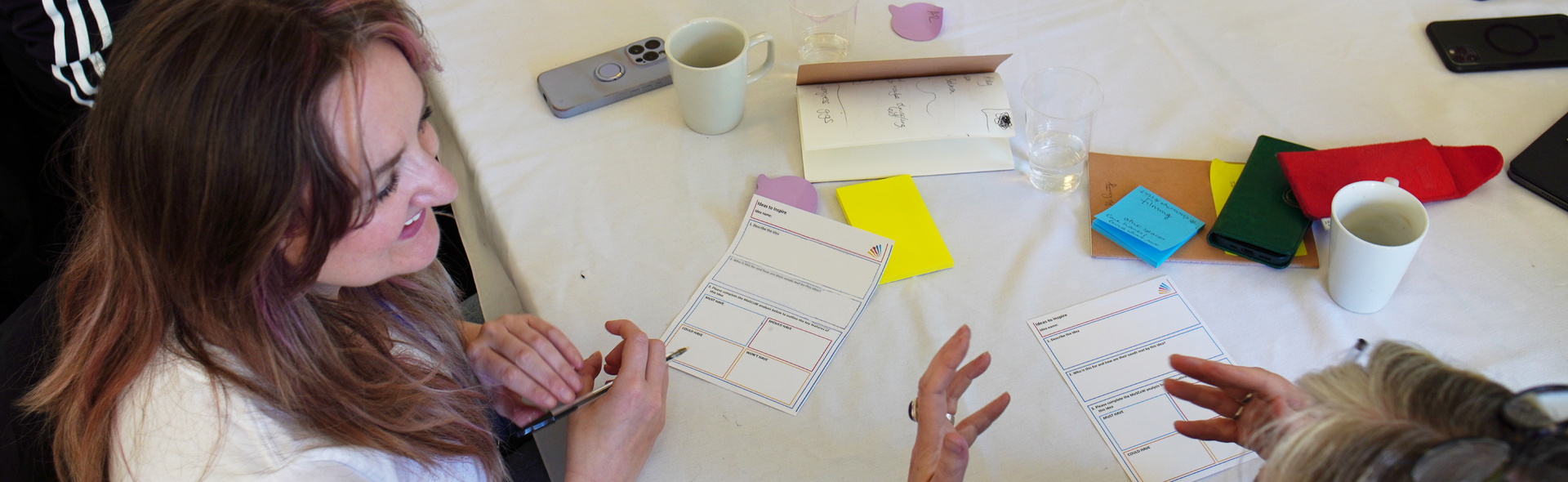 two women at a workshop smiling and talking animatedly with posits and worksheets in background