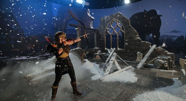 Costumed actress aims a bow in a snow-covered ruin set on Studio Ulster’s LED stage, with a large digital ogre projected on the curved screen behind her.