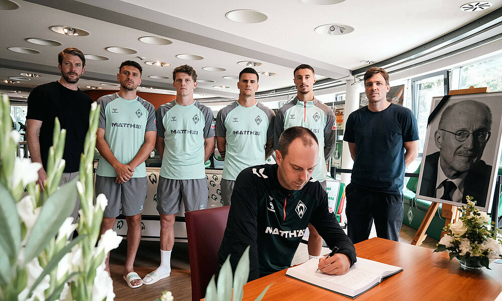 Ole Werner sitzt im Vordergrund und trägt sich in das Kondolenzbuch ein. Hinter ihm steht der Mannschaftsrat und die sportliche Leitung. 