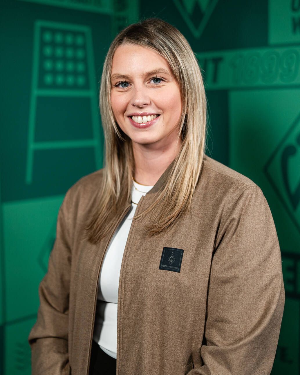 Lächelnde Frau mit langen blonden Haaren in einer braunen Jacke, vor einem grünen geometrischen Hintergrund stehend.