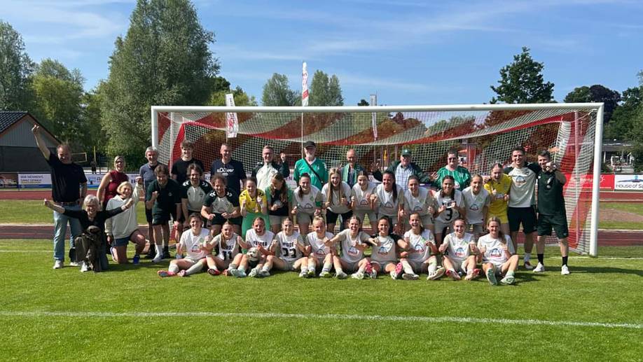Eine Fußballmannschaft posiert an einem sonnigen Tag fröhlich mit Trainern und Fans vor einem Tor.
