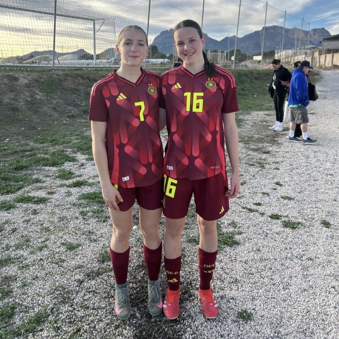 U17-Spielerinnen Catelyn Zimmermann und Matilda Schütte im DFB-Trikot vor einem Trainingsplatz.