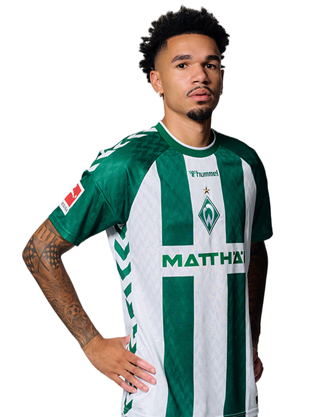Ein Mann in einem grün-weiß gestreiften Fußballtrikot von Werder Bremen steht selbstbewusst mit den Händen in den Hüften vor einem einfarbigen Hintergrund.