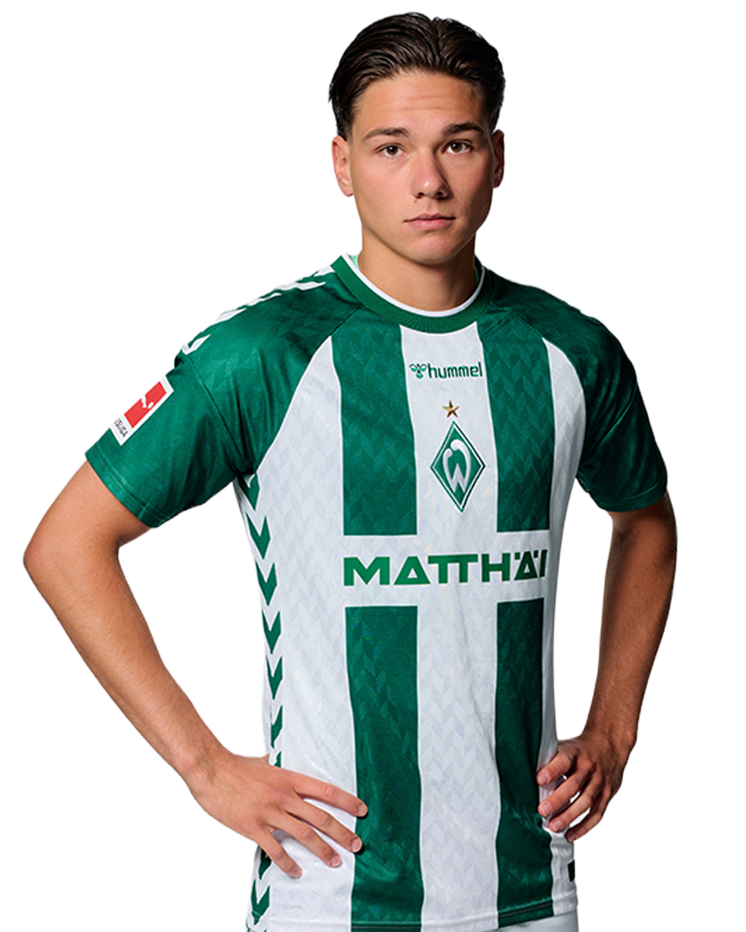 Ein Mann in einem grün-weiß gestreiften Fußballtrikot von Werder Bremen steht selbstbewusst mit den Händen in den Hüften vor einem einfarbigen Hintergrund.