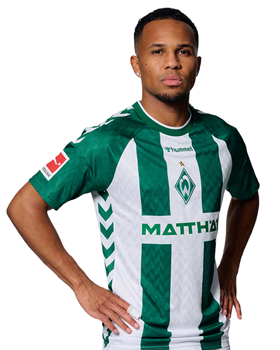 Ein Mann in einem grün-weiß gestreiften Fußballtrikot von Werder Bremen steht selbstbewusst mit den Händen in den Hüften vor einem einfarbigen Hintergrund.