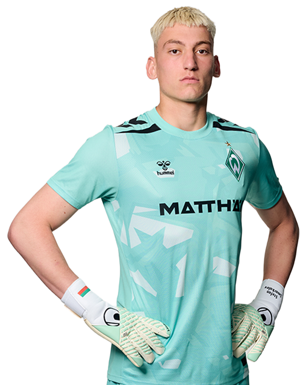Ein Mann in einem blauen Fußballtrikot von Werder Bremen steht selbstbewusst mit den Händen in den Hüften vor einem einfarbigen Hintergrund.