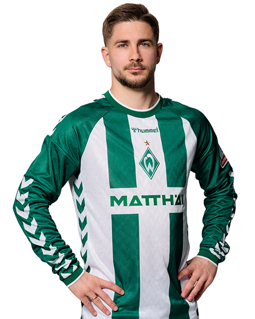 Ein Mann in einem grün-weiß gestreiften Fußballtrikot von Werder Bremen steht selbstbewusst mit den Händen in den Hüften vor einem einfarbigen Hintergrund.