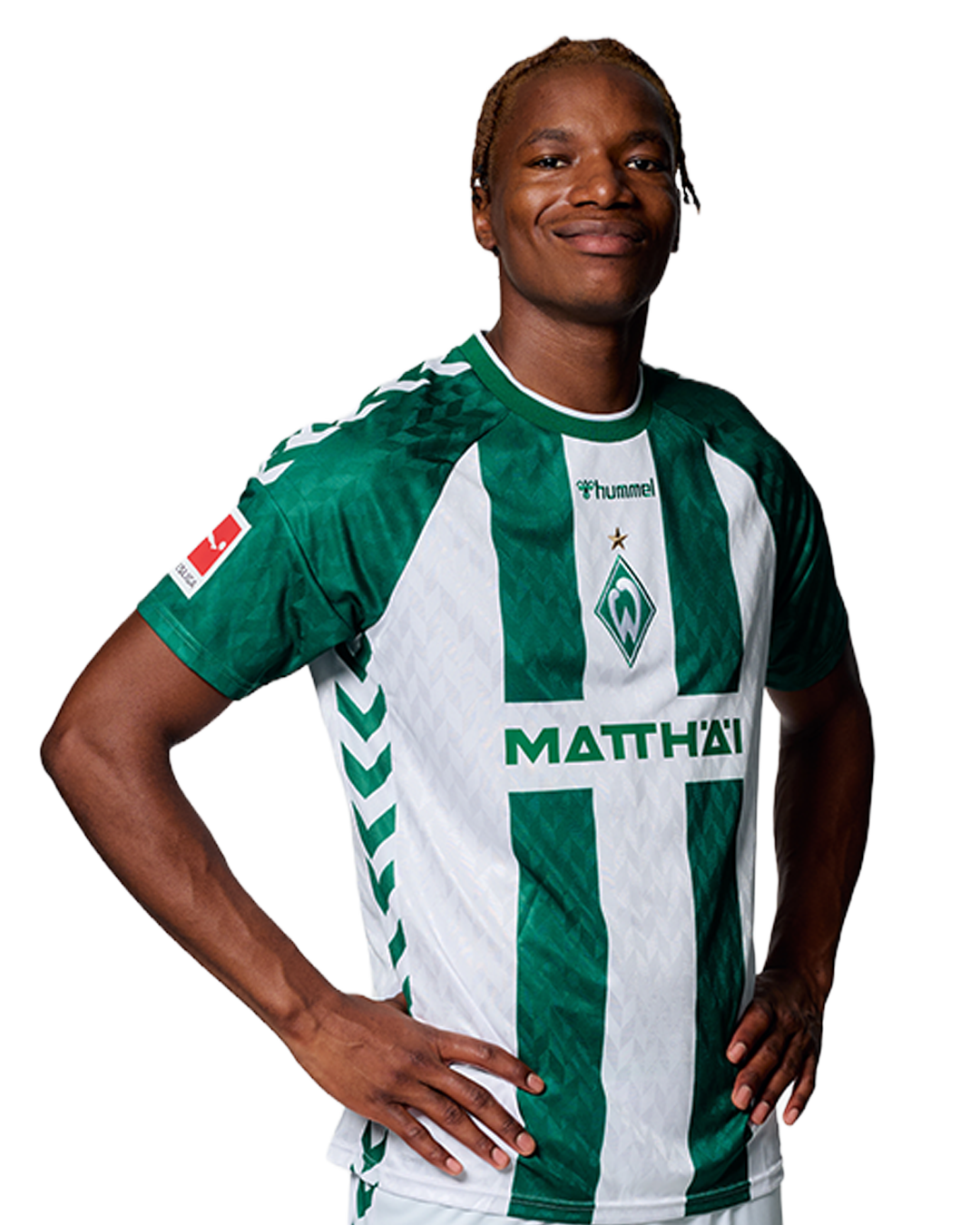 Ein Mann in einem grün-weiß gestreiften Fußballtrikot von Werder Bremen steht selbstbewusst mit den Händen in den Hüften vor einem einfarbigen Hintergrund.