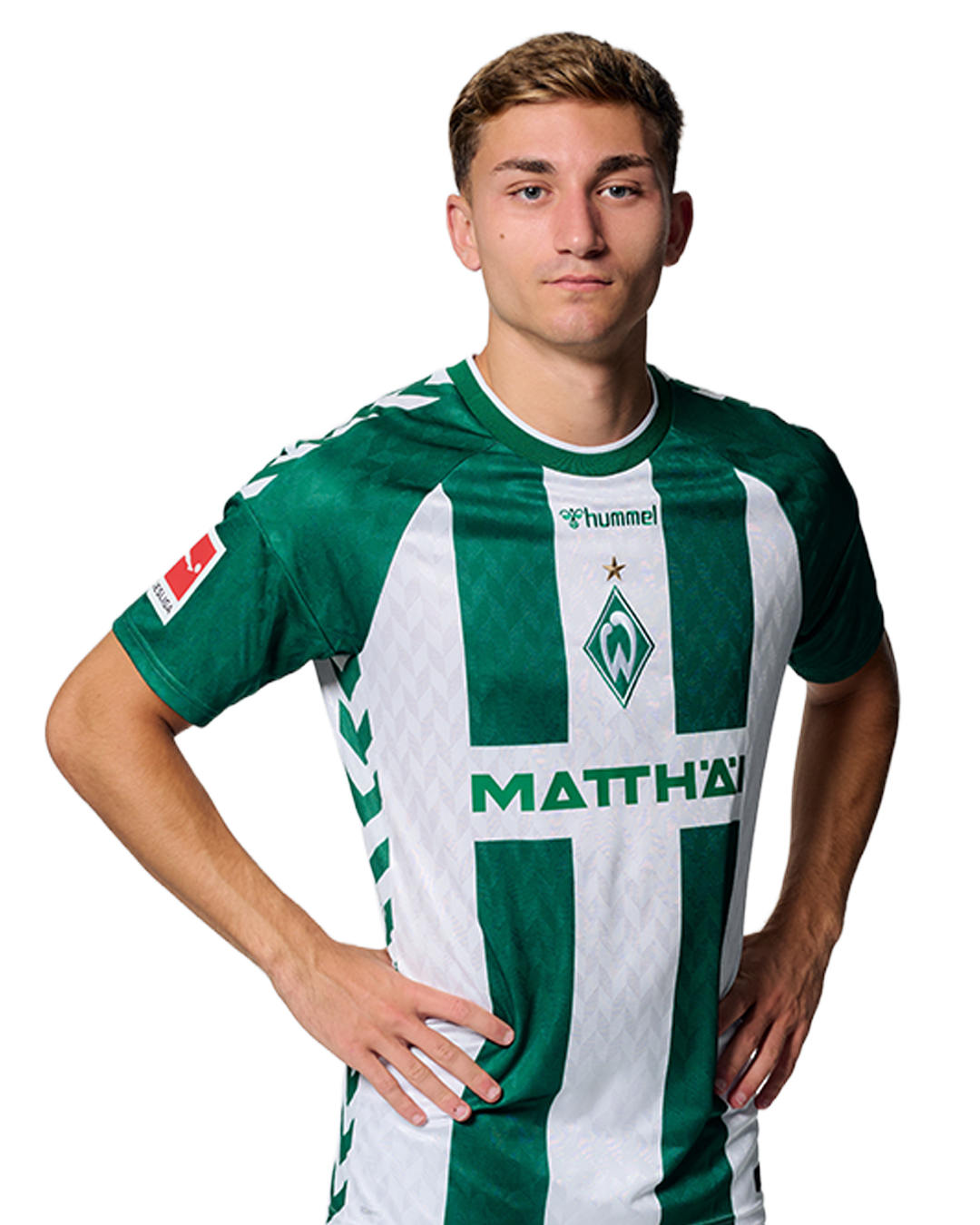 Ein Mann in einem grün-weiß gestreiften Fußballtrikot von Werder Bremen steht selbstbewusst mit den Händen in den Hüften vor einem einfarbigen Hintergrund.