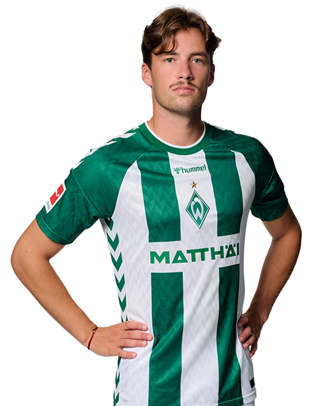 Ein Mann in einem grün-weiß gestreiften Fußballtrikot von Werder Bremen steht selbstbewusst mit den Händen in den Hüften vor einem einfarbigen Hintergrund.