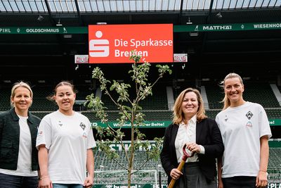 Vier Personen stehen auf einem Fußballplatz neben einem Baum. Zwei von ihnen tragen legere Sportkleidung, und im Hintergrund ist ein Schild mit der Aufschrift „Die Sparkasse Bremen“ zu sehen.