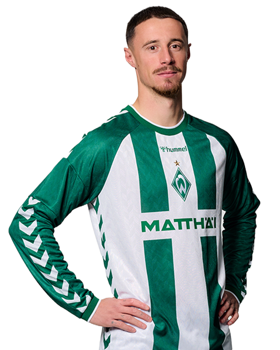 Ein Mann in einem grün-weiß gestreiften Fußballtrikot von Werder Bremen steht selbstbewusst mit den Händen in den Hüften vor einem einfarbigen Hintergrund.