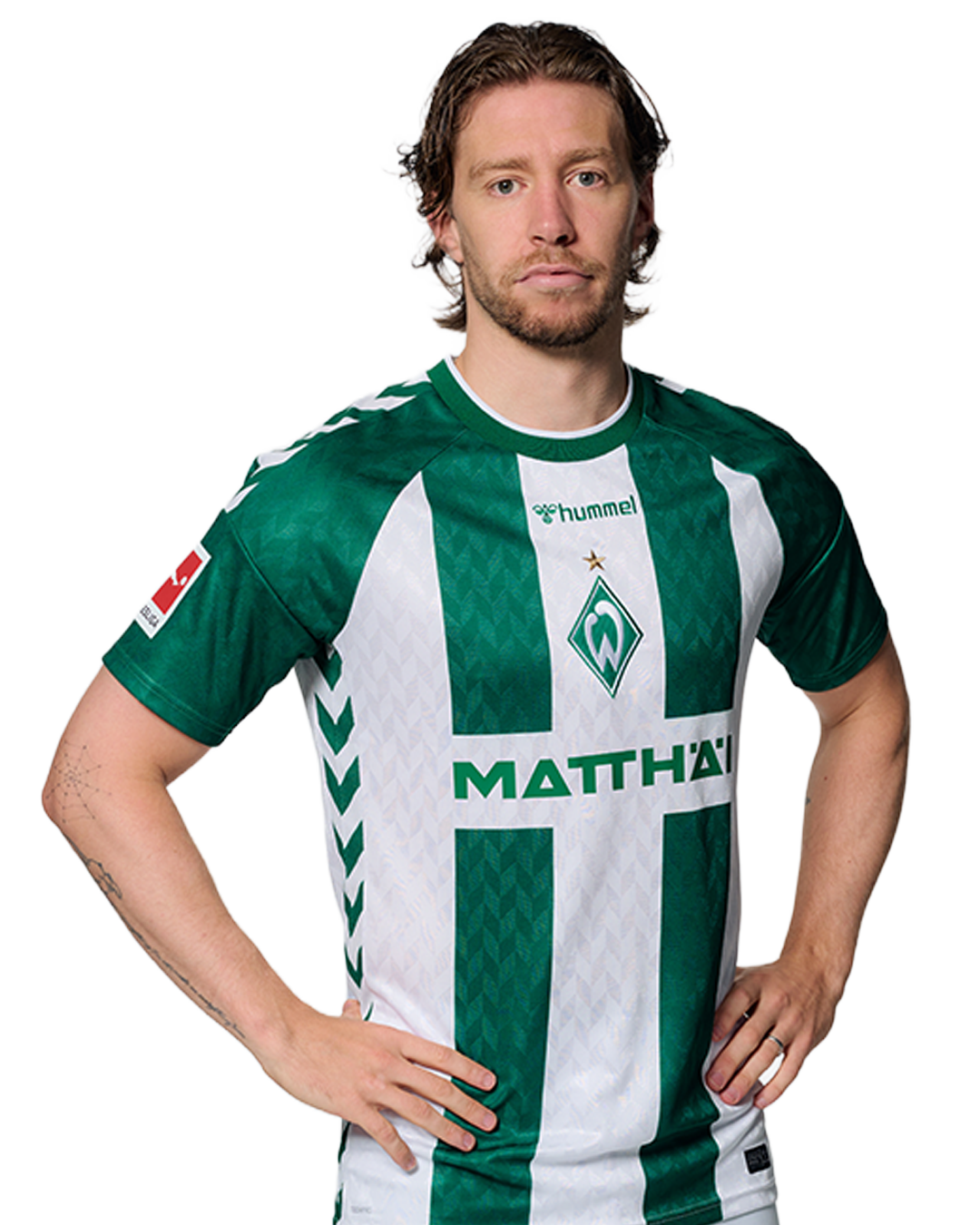 Ein Mann in einem grün-weiß gestreiften Fußballtrikot von Werder Bremen steht selbstbewusst mit den Händen in den Hüften vor einem einfarbigen Hintergrund.