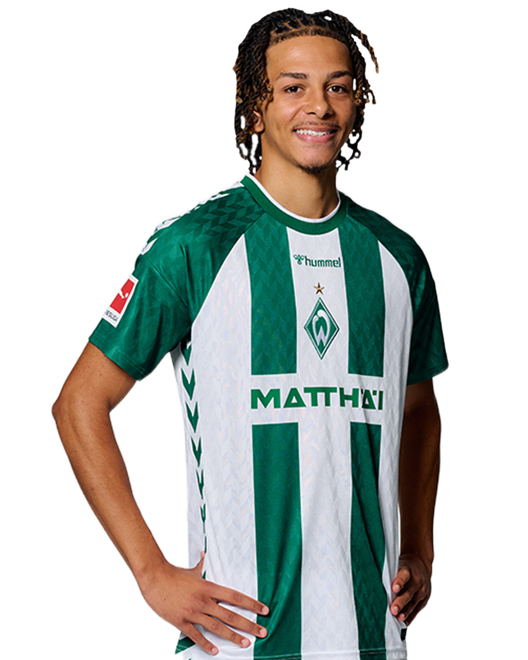 Ein Mann in einem grün-weiß gestreiften Fußballtrikot von Werder Bremen steht selbstbewusst mit den Händen in den Hüften vor einem einfarbigen Hintergrund.