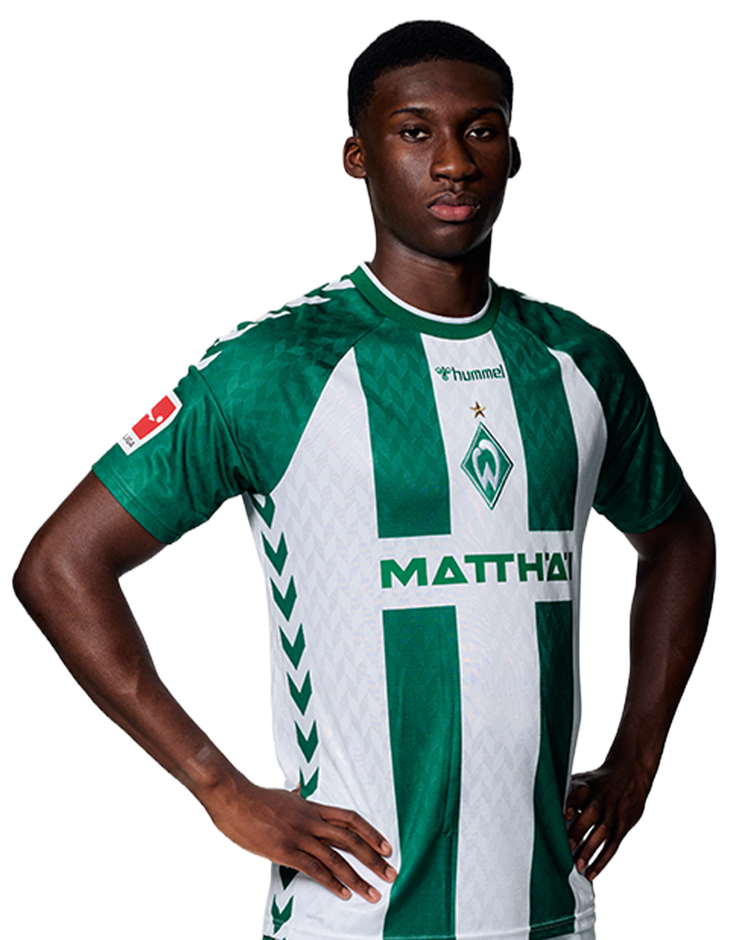 Ein Mann in einem grün-weiß gestreiften Fußballtrikot von Werder Bremen steht selbstbewusst mit den Händen in den Hüften vor einem einfarbigen Hintergrund.