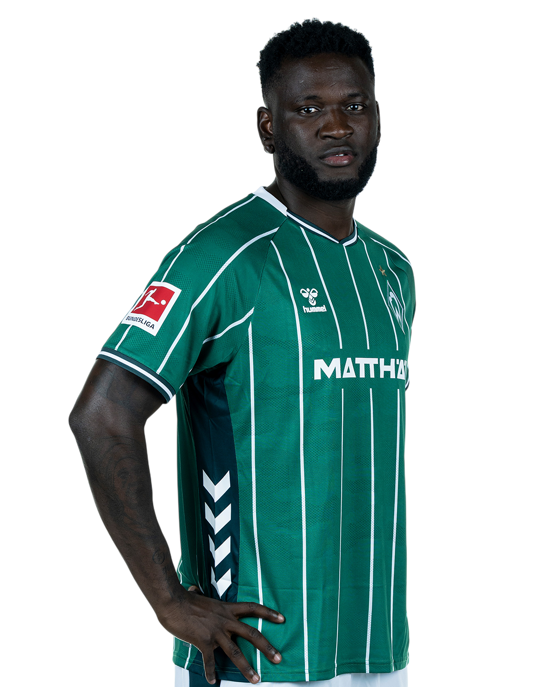 Victor Boniface in einem grün-weiß gestreiften Trikot mit der Aufschrift „MATTHÄI“ auf der Vorderseite steht mit den Händen in den Hüften da und schaut nach vorne.