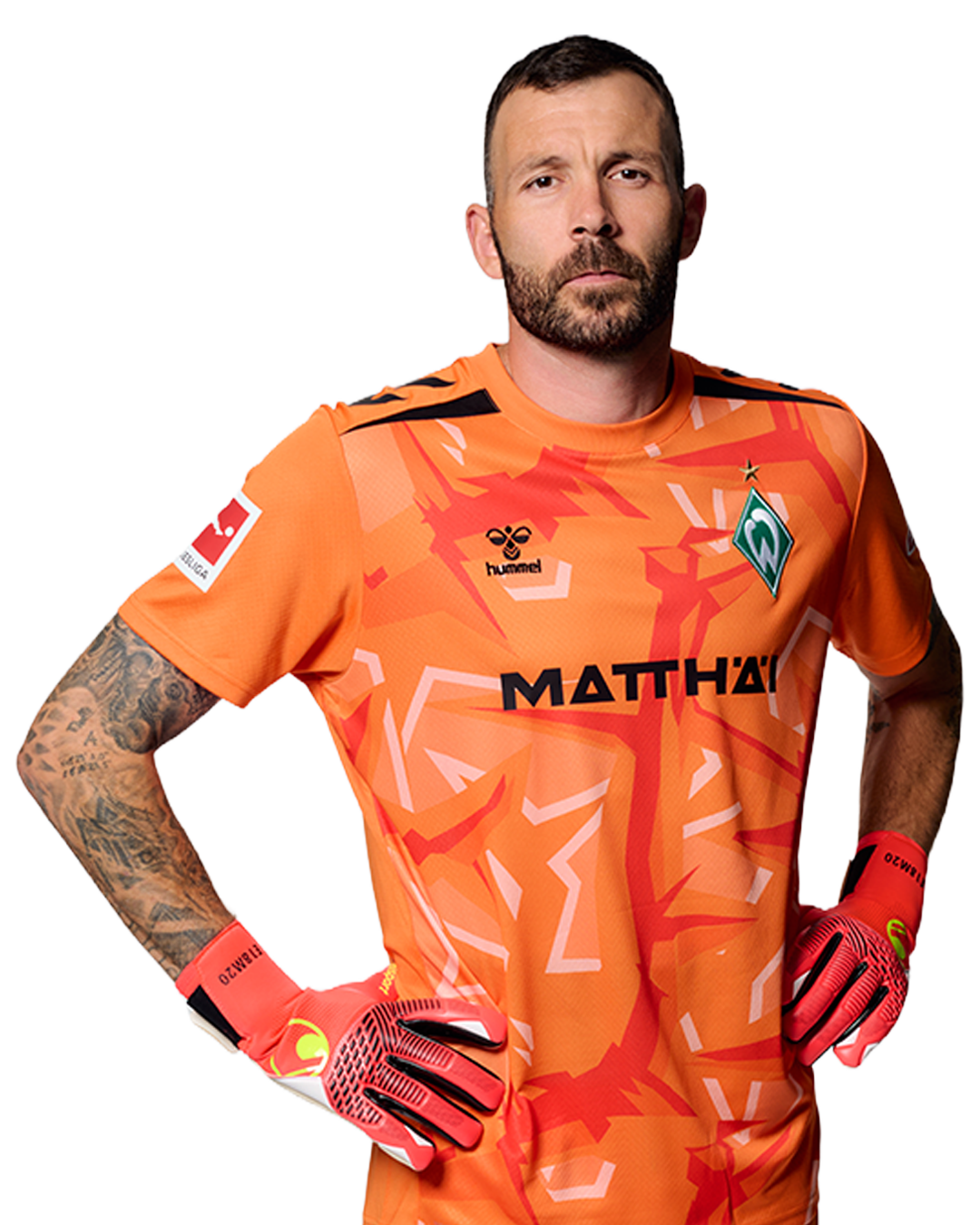 Ein Mann in einem orangenen Fußballtrikot von Werder Bremen steht selbstbewusst mit den Händen in den Hüften vor einem einfarbigen Hintergrund.