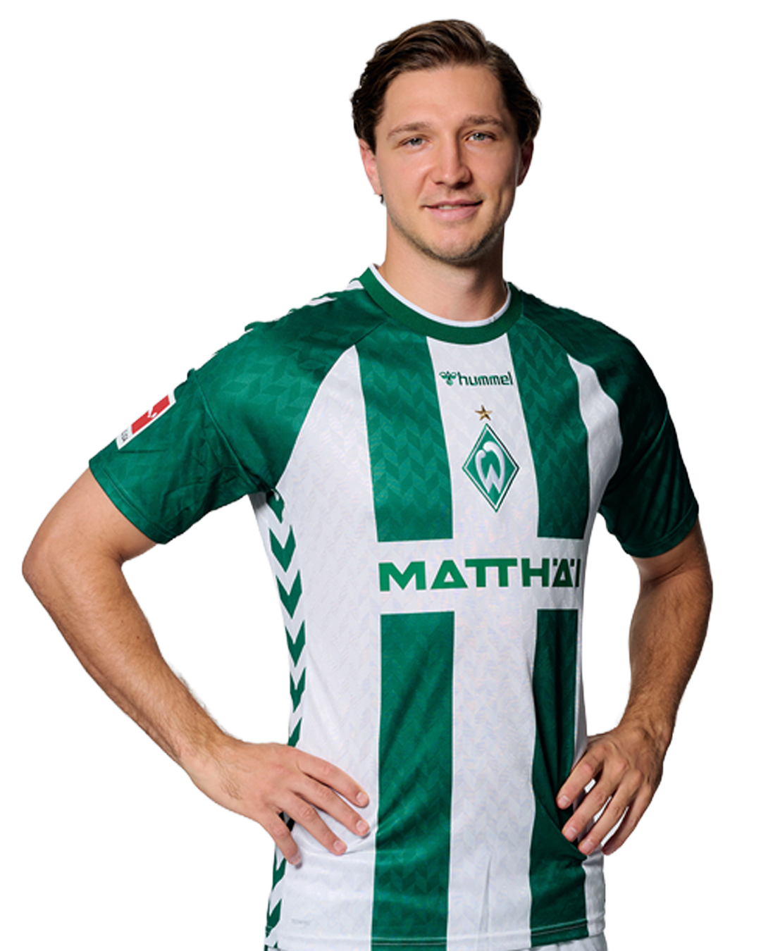 Ein Mann in einem grün-weiß gestreiften Fußballtrikot von Werder Bremen steht selbstbewusst mit den Händen in den Hüften vor einem einfarbigen Hintergrund.